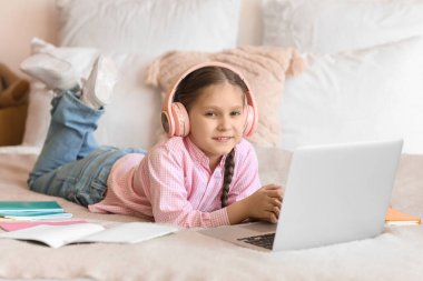 Kulaklıklı, dizüstü bilgisayarlı küçük bir kız evde yatağında bilgisayar bilimleri okuyor.