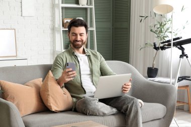 Bilgisayarlı yakışıklı adam evdeki koltukta cep telefonu kullanıyor.