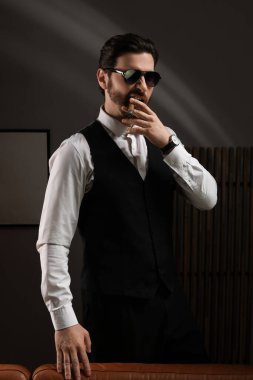 Resmi kıyafetler ve güneş gözlüğü takan yakışıklı genç adam karanlık odada kanepenin yanında puro içiyor.