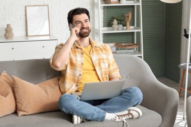 Bilgisayarlı yakışıklı adam evdeki koltukta cep telefonuyla konuşuyor.