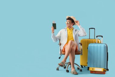 Pasaportlu ve bavullu genç işadamı mavi arka plandaki sandalyede yaz tatilini bekliyor.