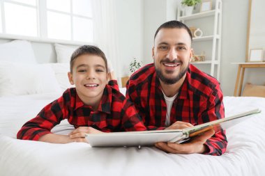 Mutlu baba ve oğlu kareli pijamalarıyla evde yatakta kitap okuyorlar.