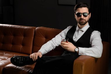 Resmi kıyafetler ve güneş gözlüğü takan yakışıklı genç adam. Karanlık odada kanepede oturmuş viski içiyor.