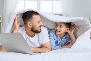 Mutlu baba ve oğlu pijamalarıyla evde dizüstü bilgisayarlarıyla yatıyorlar.