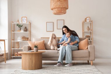 Anne, bebeği ve oyuncağıyla evdeki kanepede oturuyor.