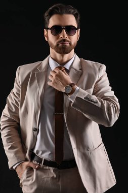 Siyah arka planda güneş gözlüklü, açık renk takım elbiseli yakışıklı bir genç.