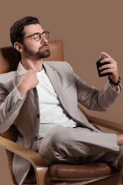 Açık renk takım elbiseli ve parfüm şişeli yakışıklı genç adam bej arka planda koltukta oturuyor.