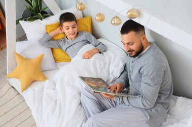 Pijamalı mutlu bir baba oğlunun yatağında kitap okuyor.