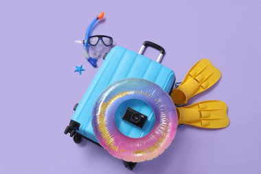 Kameralı bavul, şişme yüzük, yüzgeçli şnorkeller ve mor arka planda maske.