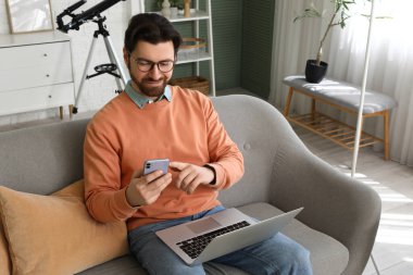 Bilgisayarlı yakışıklı adam evdeki koltukta cep telefonu kullanıyor.