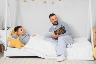 Pijamalı mutlu bir baba oğlunun yatağında kitap okuyor.