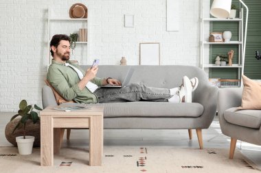 Bilgisayarlı yakışıklı adam evdeki koltukta cep telefonu kullanıyor.
