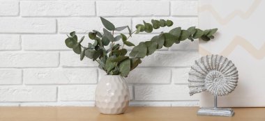 Beyaz tuğla duvarın yanındaki masada okaliptüs dalları olan heykel ve vazo.