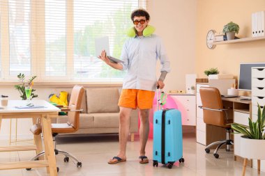 Dizüstü bilgisayarı ve bavulu olan genç işadamı yaz tatilini ofisinde bekliyor.