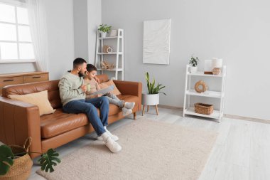 Mutlu baba ve oğlu ellerinde çay fincanıyla evdeki kahverengi deri koltukta kitap okuyorlar.