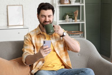 Evdeki koltukta cep telefonu kullanarak çay içen yakışıklı bir adam.