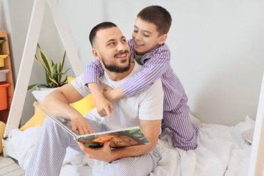 Mutlu baba ve oğlu pijamalarıyla evde kitap okuyorlar.