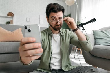 Gözlüklü düşünceli genç adam evde cep telefonu kullanıyor.