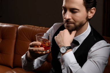 Kol saati ve bir bardak viskisi olan yakışıklı genç adam karanlık odada kanepede oturuyor.