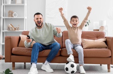 Mutlu baba ve oğlu futbol topuyla evdeki kahverengi deri koltukta futbol izliyorlar.