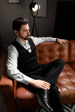 Resmi giyinmiş yakışıklı genç adam karanlık odada kanepede oturuyor.