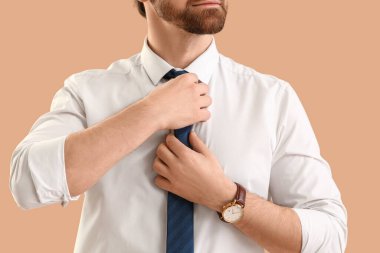 Resmi giyinmiş yakışıklı genç adam bej arka planda kravatını düzeltiyor.