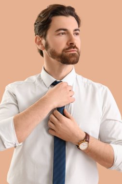Resmi giyinmiş yakışıklı genç adam bej arka planda kravatını düzeltiyor.