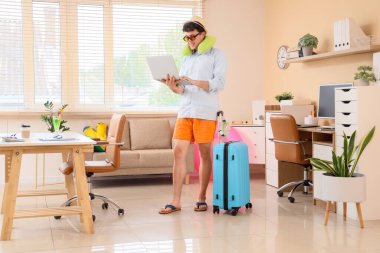 Genç iş adamı seyahat aksesuarlarıyla ofiste dizüstü bilgisayar kullanıyor. Yaz tatili