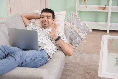 Dizüstü bilgisayarlı genç adam evdeki kanepede sohbet ediyor.