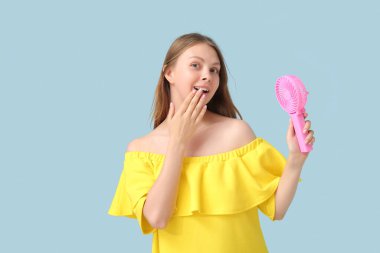 Mavi arka planda mini vantilatörü olan şaşırmış genç bir kadın.