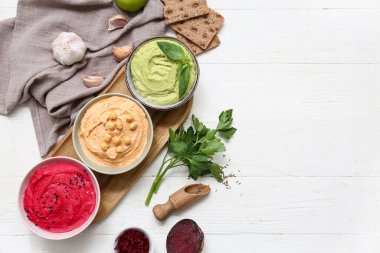 Beyaz ahşap arka planda sarımsak, maydanoz, pancar ve kraker içeren farklı lezzetli humus kaseleri.
