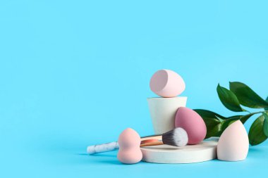 Mavi arka planda makyaj süngerleri, fırçaları ve taze yaprakları olan dekoratif podyumlar