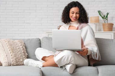 Evdeki kanepede oturan rahat battaniyeli ve laptoplu güzel Afro-Amerikalı kadın.