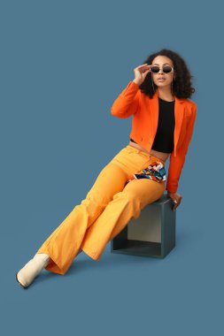 Turuncu ceketli, mavi arka planda oturan Afro-Amerikan bir kadın.