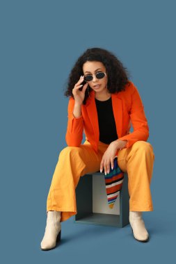 Turuncu ceketli, mavi arka planda oturan Afro-Amerikan bir kadın.