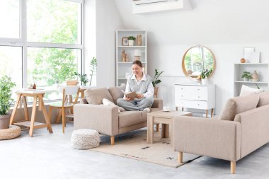 Oturma odasındaki gri koltukta kitap okuyan gülümseyen genç kadın.