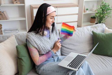LGBT bayrağı ve modern dizüstü bilgisayarıyla oturma odasındaki koltukta oturan genç ve güzel bir kadın.