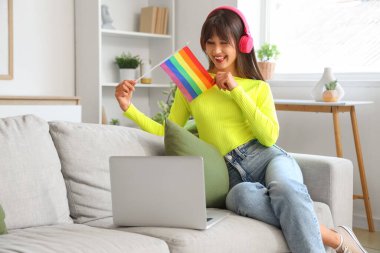 Kulaklıklı, LGBT bayrağı ve oturma odasındaki kanepede oturan modern dizüstü bilgisayarlı güzel bir kadın.