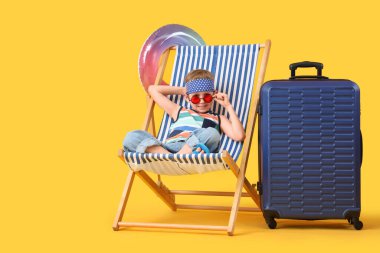 Sarı arka planda şezlongda oturan şişme yüzüklü ve valizli şirin küçük çocuk. Seyahat kavramı