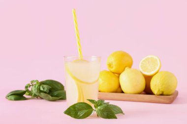Bir bardak taze limonata, fesleğen yaprakları ve pembe arka planda limonlar.