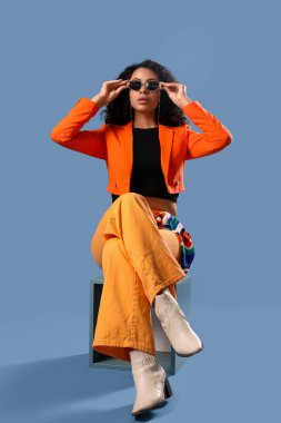 Turuncu ceketli, mavi arka planda oturan Afro-Amerikan bir kadın.