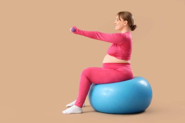 Sportif, genç, hamile kadın, kahverengi arka planda halterli halterlerle antrenman yapıyor.