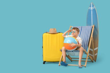 Güverte sandalyesinde oturan şirin küçük çocuk şişme top, bavul ve mavi arka planda sörf tahtasıyla. Seyahat kavramı