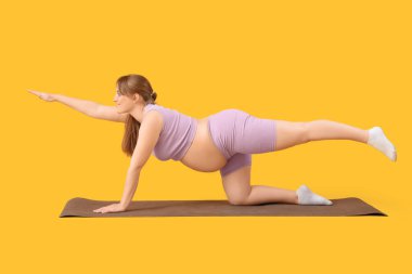 Sportif genç hamile kadın sarı arka planda yoga minderi eğitimi alıyor.