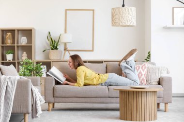 Gülümseyen genç kadın oturma odasındaki gri koltukta dergi okuyor.
