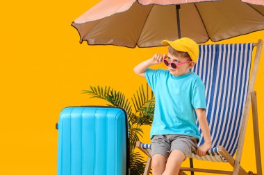 Sarı arka planda bavulu ve şemsiyesiyle güverte koltuğunda oturan sevimli küçük çocuk. Seyahat kavramı