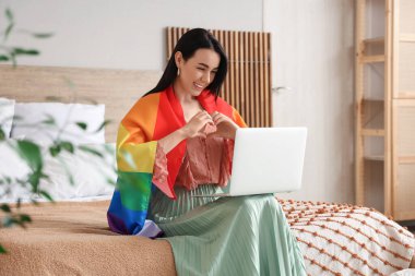 LGBT bayrağı ve modern dizüstü bilgisayarı olan güzel genç bir kadın kalp hareketi gösteriyor ve yatak odasında oturuyor.