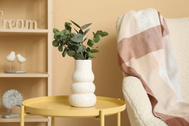 Oturma odasındaki bej duvarın yanındaki masada okaliptüs dalları olan vazo, yakın plan.