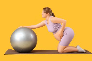Sportif genç hamile kadın, sarı arka planda yoga minderinde fitball antrenmanı yapıyor.