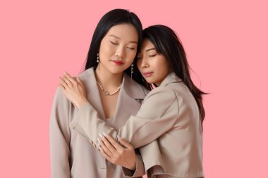 Güzel, genç, mutlu Asyalı kız kardeşler şık bej kıyafetler içinde pembe arka planda sarılıyorlar.